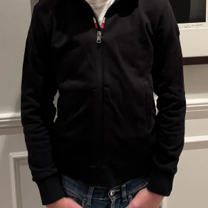 Svart cardigan/ siptröja från Moncler - Säljer en snygg svart cardigan från Moncler i mycket bra skick. Den har en dragkedja framtill och Monclers logga på ärmen. Cardiganen är långärmad och gjord i ett bekvämt material. Modellen på bilden är 168cm och 52kg, var inte rädd att höra av dig vid intresse samt frågor, pris är ej hugget i sten😉🤩