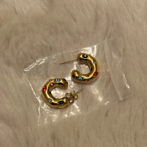 Guldörhängen med färgglada stenar - Säljer ett par snygga guldörhängen med coola färgglada stenar. De har en unik, böjd form som verkligen sticker ut. Perfekta för att liva upp vilken outfit som helst! 
