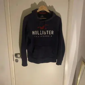 Säljer en snygg och bekväm mörkblå hoodie från Hollister. Den har en stor logga på framsidan och en praktisk magficka. Perfekt för kyliga dagar eller mysiga hemmakvällar. Passar både till vardags och avslappnade tillfällen.