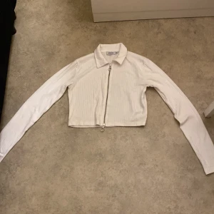 Vit croppad jacka från Junkyard - Säljer en snygg vit croppad jacka från Junkyard i storlek M. Jackan har en dragkedja framtill och långa ärmar. Perfekt för vår och sommar, och funkar både till vardags och fest. Den är i bra skick och redo för en ny ägare!