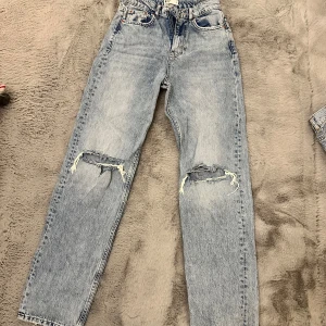 Blå jeans med slitna detaljer - Säljer ett par snygga blå jeans från Gina Tricot i storlek 32. De har en cool slitning på knäna och en klassisk femficksdesign. Perfekta för en avslappnad stil och passar till alla tillfällen. Dragkedja och knapp framtill.