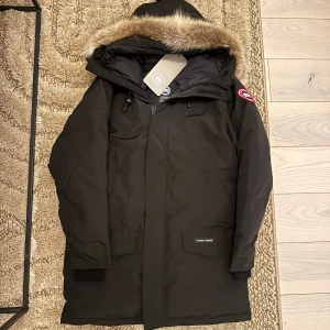 Svart parkajacka från Canada Goose - Säljer en svart parkajacka från Canada Goose i nytt skick. Jackan har en avtagbar huva med pälsdetaljer och en dragkedja framtill. Perfekt för kalla vinterdagar med sin varma och skyddande design. Märkeslogga på ärmen ger en snygg touch. Passar både till vardags och mer formella tillfällen. Jackan är i storlek S men passar även M då modellen är lång i sig. Har kvar lapp på jackan. Finns inget kvitto då det inte är äkt*. Men om du kan jackor och kvalite går det att se att allt är felfritt.