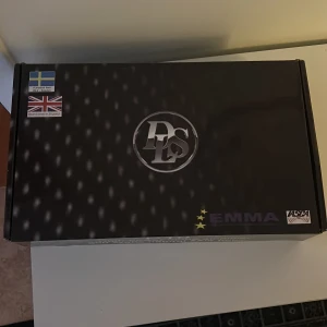 DLS EMMA slutsteg Produktförpackning  - Det här är en produktförpackning från DLS med EMMA-märkning. Förpackningen är svart med logotyper och flaggor från Sverige och Storbritannien. Inuti finns det skyddande material och dokumentation. Perfekt för den som söker högkvalitativa ljudprodukter. Oanvänd och oöppnad.