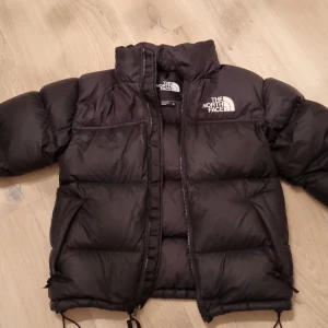 Svart dunjacka från The North Face - Säljer en svart dunjacka från The North Face i mycket bra skick. Jackan är perfekt för kalla vinterdagar och har en dragkedja framtill. Den är varm och bekväm med en klassisk pufferstil. Passar perfekt för både vardag och utomhusaktiviteter.
