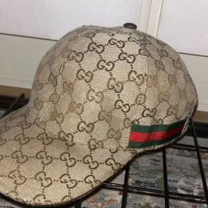 Beige keps från Gucci - Säljer en snygg beige keps från Gucci med deras ikoniska GG-mönster. Kepsen har en klassisk design med en röd och grön randdetalj på sidan. Perfekt för att ge din outfit en lyxig touch. Tillverkad i Italien och i mycket bra skick.