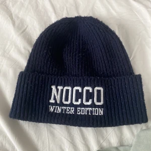 Nocco Mössa - Säljer en mörkblå stickad mössa från NOCCO, Winter Edition. Den är ribbad och har en broderad text framtill. Perfekt för kalla vinterdagar och håller dig varm och stilren. Passar de flesta huvudstorlekar.