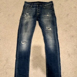 Blå slitna jeans från Jack & Jones - Säljer ett par blå slitna jeans från Jack & Jones i modellen Slim/Glenn. De har en cool distressed look med flera revor och slitningar. Perfekta för en avslappnad och trendig stil. Passar bra till både vardag och fest!
