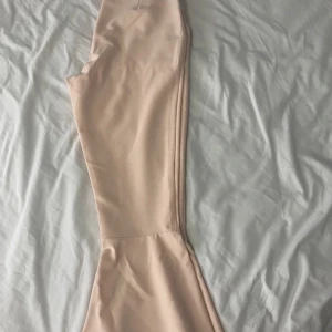 Beige byxor med utsvängda ben - Säljer ett par snygga beige/gammelrosa byxor med utsvängda ben. De är högmidjade och har en stilren design som passar perfekt till både vardag och fest. Materialet känns mjukt och bekvämt, vilket gör dem sköna att bära hela dagen. Perfekta för dig som vill ha en trendig look!