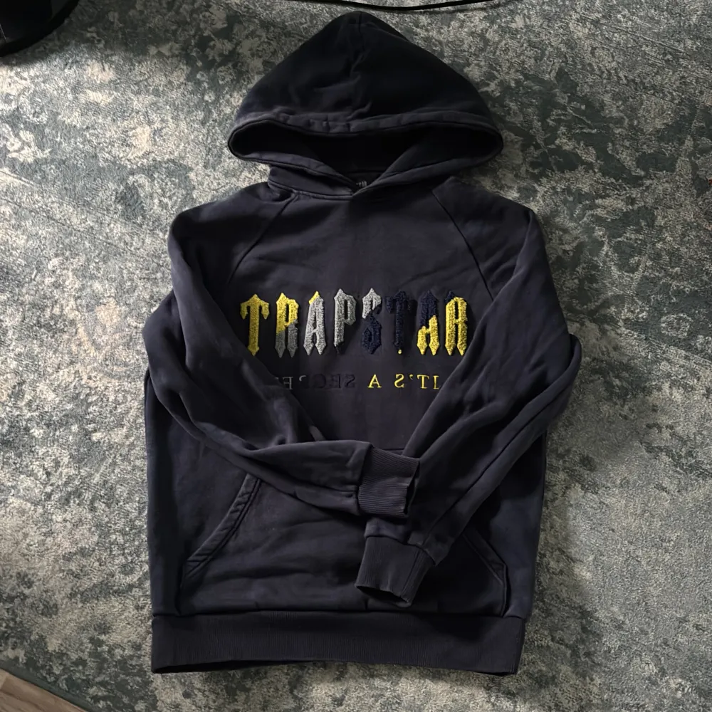 Säljer en snygg svart hoodie från Trapstar med coolt tryck i gult och vitt på framsidan. Den har en klassisk känguruficka och en bekväm huva. Perfekt för en avslappnad stil eller streetwear-look. Passar både till vardags och chillkvällar.. Tröjor & Koftor.