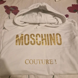 Vit hoodie från Moschino - Säljer en snygg vit hoodie från Moschino med guldtryck. Den har en klassisk design med huva och dragsko. Perfekt för en stilren och bekväm look. Passar både till vardags och festliga tillfällen.