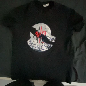 Svart t-shirt med tryck - Säljer en svart moncler t-shirt med ett coolt tryck i rött och vitt. Perfekt för en avslappnad stil eller att bära på fest. T-shirten är i bra skick och har en normal passform. Grym att matcha med jeans eller shorts den har andvänts ett tag kanske över 4 månader. Köpt från trendtorget för 2000 , jag säljer den för 500