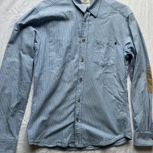 Blå randig skjorta från Selected Jeans - Säljer en snygg blå randig skjorta från Selected Jeans. Skjortan har långa ärmar och bruna knappar, samt stiliga armbågslappar i beige. Perfekt för vardag!