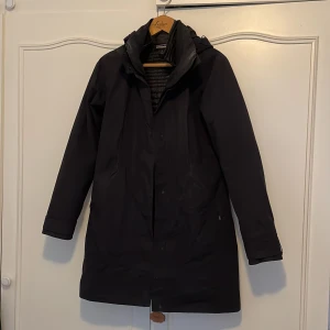 Svart kappa från Patagonia - Säljer en stilren svart kappa från Patagonia i storlek M. Jackan har långa ärmar och en praktisk huva, perfekt för kyliga dagar. Den är i  okej skick och en ficka har ett hål som behöver repareras.  