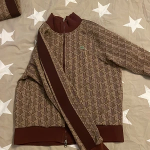 Lacoste track suit - Säljer en snygg brun mönstrad jacka från Lacoste i mycket bra skick. Jackan har ett coolt geometriskt mönster och den klassiska gröna krokodilen på bröstet. Den har dragkedja och är långärmad, perfekt för höst och vår. Passformen är normal och materialet känns bekvämt. Perfekt för dig som vill ha en stilren look! Track suit med byxor. 