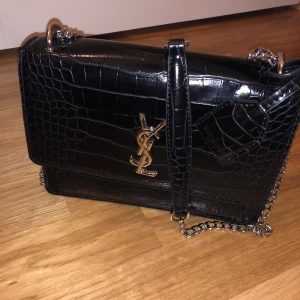 Svart axelväska från YSL - Säljer en elegant svart axelväska från YSL i krokodilpräglat skinn. Väskan har en kedjerem och ett stilrent YSL-emblem i guld på framsidan. Perfekt för både vardag och fest! 🖤