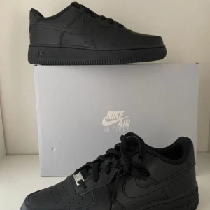 Svarta Nike Air Force sneakers - Säljer ett par klassiska svarta Nike Air Force sneakers i nytt skick. De är helt svarta med snörning och har den ikoniska Nike-loggan på sidan. Perfekta för både vardag och fest! Kommer i originalkartong.