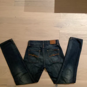 Nudie jeans - Säljer ett par mörkblå jeans i bra skick. De har en snygg slitningseffekt på framsidan och baksidan, vilket ger dem en cool look. Jeansen har en klassisk femficksdesign och knappar i midjan. Perfekta för både vardag och fest!