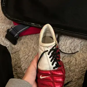 Säljer en cool röd och vit fotbollssko från Nike. Skon har en snygg design med svarta snören och den klassiska Nike-loggan på sidan. Perfekt för fotbollsträning eller match. Skon är i bra skick och redo för nya äventyr på planen!