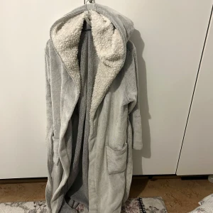 Grå fleece morgonrock från Loving Loungewear - Mysig grå morgonrock i fleece från Loving Loungewear. Den har en fluffig insida och en praktisk huva för extra värme. Perfekt för kalla morgnar eller mysiga kvällar hemma. Rocken har ett bälte i midjan och två fickor framtill.