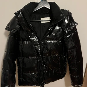 Svart pufferjacka från Calvin Klein - Säljer en snygg svart pufferjacka från Calvin Klein. Den är glansig och har en dragkedja framtill. Perfekt för kalla dagar med sin varma vaddering. Jackan har en huva och långa ärmar, den är jätte varm, bara använt en vinter så den har inga defekter, är som ny! Nypris 2000kr