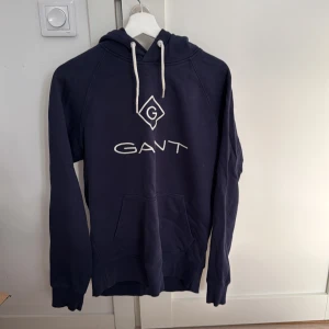 Mörkblå hoodie från GANT - Säljer en snygg mörkblå hoodie från GANT i storlek M. Den har en klassisk design med vit logga på bröstet och justerbara snören i huvan. Perfekt för en avslappnad stil och passar bra till både jeans och joggers. Använd men i bra skick!
