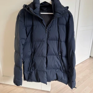 Svart pufferjacka från J.Lindeberg - Säljer en snygg svart pufferjacka från J.Lindeberg. Den är perfekt för kalla dagar med sin quiltade design och dragkedja framtill. Jackan har en huva och två praktiska fickor med dragkedjor. Den är i mycket bra skick och passar perfekt för vintern. Perfekt för dig som vill hålla stilen i kylan! Storlek man medium.