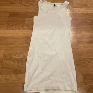 Vit ribbad klänning - Säljer en vit ribbad klänning i ett enkelt och stilrent snitt. Klänningen är ärmlös och har en tight passform som framhäver figuren. Perfekt för sommardagar eller som ett basplagg att styla med olika accessoarer. Observera att det finns en liten fläck på framsidan.