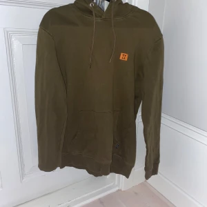 Olivgrön hoodie med logga - Säljer en olivgrön hoodie i bra skick med en liten orange logga på bröstet. Den har en klassisk känguruficka och justerbar huva med snören. Perfekt för höst och vinter, superbekväm och stilren. Passar både till vardags och chillkvällar. 🧡