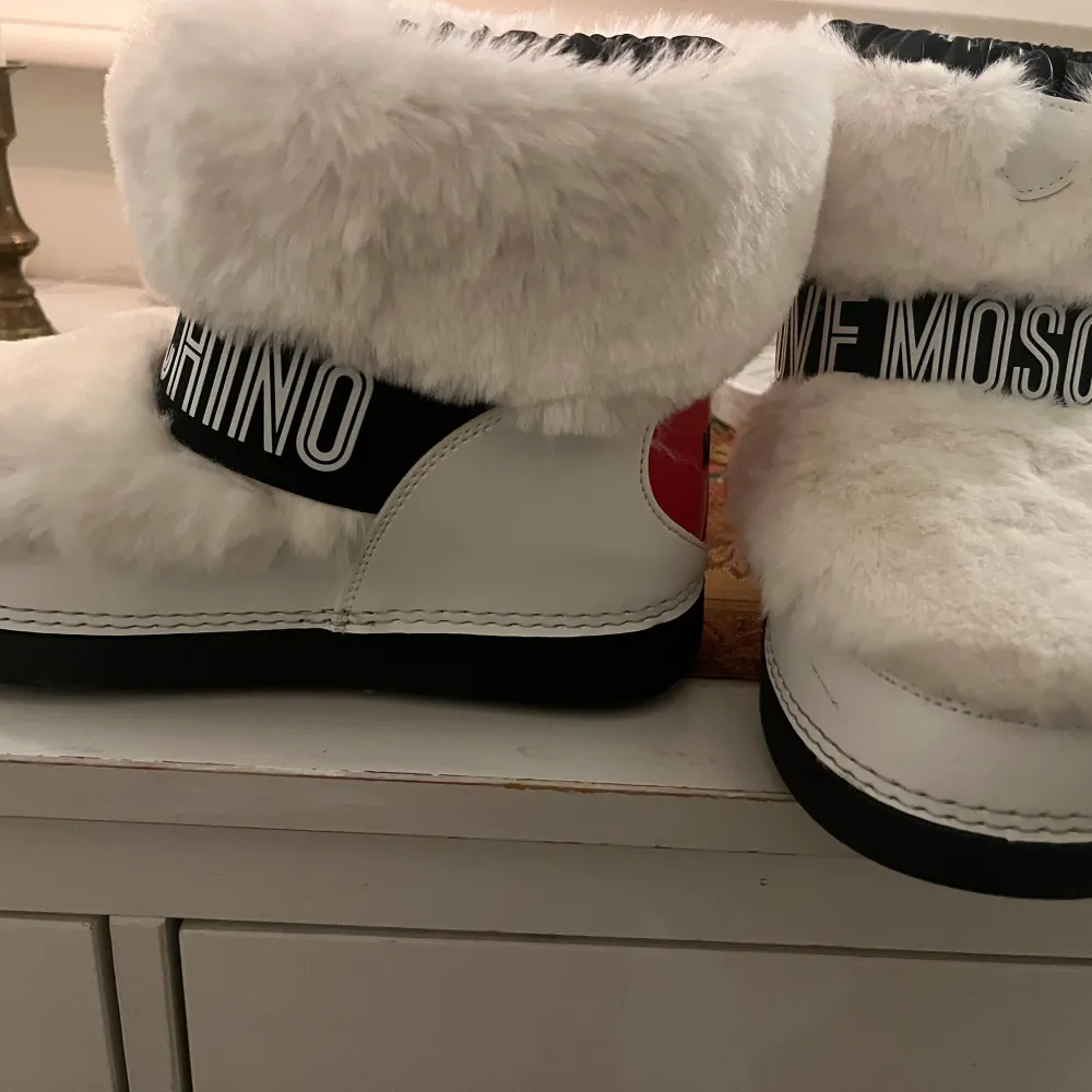 Superfina vita boots från Love Moschino med fluffig päls och svart textband runt skaftet. De har en cool design med ett rött hjärta på baksidan och är perfekta för vintern. Insidan är fodrad för extra värme och komfort. Perfekta för att hålla stilen även när det är kallt ute!. Skor.