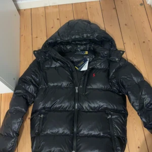 Polo Ralph Lauren jacka - Säljer en snygg svart dunjacka från Polo Ralph Lauren i storlek S. Jackan har en quiltad design med dragkedja och en liten röd logga på bröstet. Perfekt för kalla vinterdagar med sin varma och bekväma passform. Prislapp finns