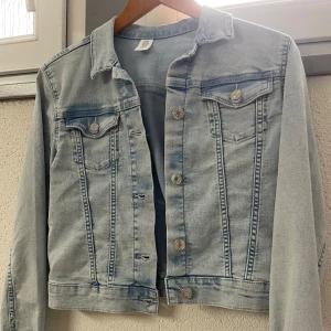 Ljusblå jeansjacka från &Denim - Säljer en snygg ljusblå jeansjacka från &Denim. Jackan har en klassisk design med knappar framtill och två bröstfickor med knappstängning. Perfekt för vår och sommar!