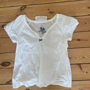 Vit blus från Odd Molly - Säljer en superfin vit blus från Odd Molly. Den har en söt liten rosett framtill och ett broderat märke på baksidan. Blusen är kortärmad och har en v-ringad hals. Perfekt för en avslappnad sommarlook! Blusen är använd två gånger. ses skår storlek 0 i vilken typ betyder xs.