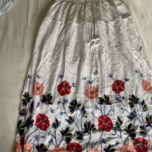 Vit långkjol med broderade blommor - Säljer en superfin vit långkjol med färgglada broderade blommor. Kjolen har en elastisk midja med knytband för perfekt passform. Den är perfekt för sommarens alla fester och ger en bohemisk touch till din outfit. Materialet känns lätt och luftigt, vilket gör den idealisk för varma dagar.