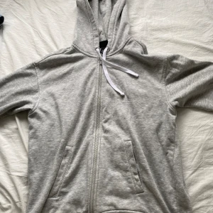Grå hoodie med dragkedja - Säljer en enkel och bekväm grå hoodie med dragkedja. Perfekt för vardagsbruk eller när du bara vill mysa hemma. Den har en justerbar huva med vita snören och praktiska fickor framtill. Passar bra till både jeans och mjukisbyxor!