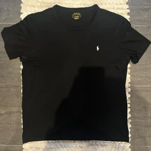 Svart t-shirt från Ralph Lauren - Säljer en klassisk svart t-shirt från Ralph Lauren. Den har en liten vit broderad logga på bröstet och är gjord i mjuk bomull. Perfekt för en stilren och avslappnad look. Passar till både vardag och fest!