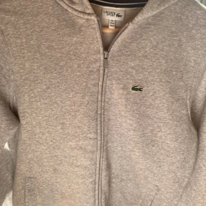 Grå hoodie från Lacoste - Säljer en snygg och bekväm grå hoodie från Lacoste. Den har en dragkedja framtill och den klassiska krokodilloggan på bröstet. Den är i bra begagnat skick (lite nåpprig) hel och ren. Den är liten i storleken så den passar dig som är runt 170💯💯