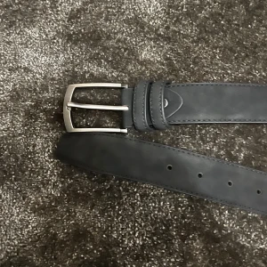 Svart skärp i skinn - Säljer ett stilrent svart skärp i skinn och mocka med ett klassiskt silverfärgat spänne. Perfekt för både vardag och fest! Skärpet har flera hål för justerbar passform och är i nyskick.