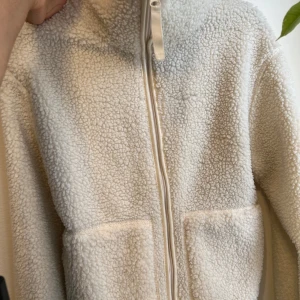 Vit teddyjacka med dragkedja - Säljer en supermysig vit teddyjacka med dragkedja framtill. Perfekt för kyliga dagar när du vill vara både varm och stilren. Jackan har en enkel design med två fickor fram och en hög krage.