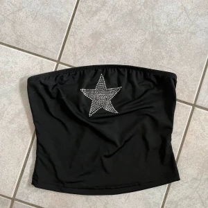 Svart tubtopp med stjärndetalj - Snygg svart tubtopp med en glittrig stjärna framtill. Perfekt för festliga tillfällen eller en utekväll. Toppen är axelbandslös och har en tight passform som sitter bra på plats. skriv vid frågor💗