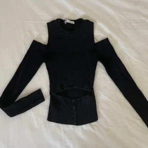 Svart ribbad topp från Zara - Säljer en svart ribbad topp från Zara. Den har en unik design med cut-out detaljer vid axlarna och knappar framtill. Perfekt för en trendig look!