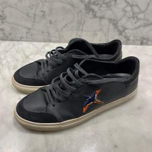 Säljer ett par svarta sneakers från Axel Arigato i storlek 43. Skorna har en cool design med en broderad fågel i orange och blått på sidan. De är gjorda i läder med mockadetaljer på tån och har en vit sula. 