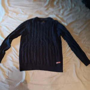 Mörkblå stickad tröja - Säljer en snygg mörkblå stickad tröja med kabelstickat mönster. Perfekt för kyliga dagar och passar både till vardags och lite finare tillfällen. Tröjan har långa ärmar och en klassisk rund halsringning. Supermysig och stilren!