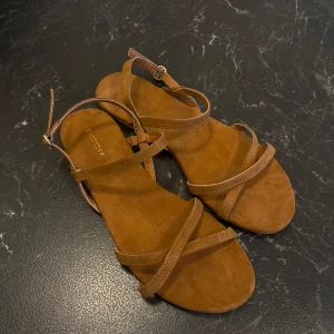 Bruna sandaler - Snygga bruna sandaler från H&M i storlek 40. Smala remmar och en justerbar spänne runt ankeln. Perfekta för sommardagar och ger en stilren look till vilken outfit som helst. Använda men i bra skick.
