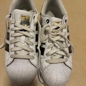 Adidas Superstar i stl 38,5 i fint skick - Adidas Superstar i vit, svart och guld i stl 38,5 i fint använt skick. Inga direkta slitningar el missfärgningar