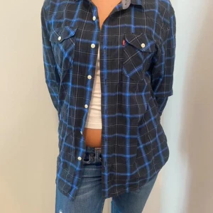 Svart och blå rutig skjorta från Levi's - Säljer en snygg svart och blå rutig skjorta från Levi's. Skjortan har långa ärmar och knappar framtill. Perfekt för en avslappnad stil eller en dag på stan. Den är i bra skick och redo för nya äventyr! 🖤💙