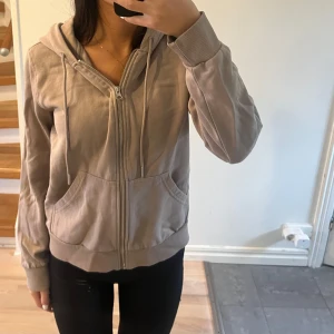 Beige huvtröja med dragkedja - Säljer en mysig beige-lila huvtröja med dragkedja framtill. Tröjan har långa ärmar och en praktisk huva med snören. Perfekt för höst och vår, och passar till vardags. Storleken är normal och materialet känns mjukt och bekvämt.