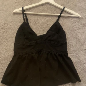 Svart topp med spetsdetaljer - Snygg svart topp med smala axelband och spetsdetaljer. Den har en v-ringad design och är perfekt för sommarens fester. Materialet är lätt och luftigt, vilket gör den bekväm att bära. 