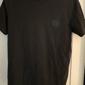 Svart t-shirt från Moncler - Säljer en stilren svart t-shirt från Moncler i storlek S. Den har en diskret logga på bröstet och är perfekt för både vardag och fest. Materialet känns mjukt och skönt mot huden. Passar perfekt till ett par jeans nu på hösten. Hör av dig vid minsta fundering👑