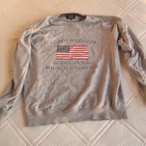 Säljer en grå sweatshirt från Vinson Polo Club med ett tryck av en amerikansk flagga och texten 'INTERNATIONAL POLO CLUB HERITAGE' på framsidan. Tröjan är långärmad och har en klassisk rund halsringning. Perfekt till hösten är för den är inte sö varm