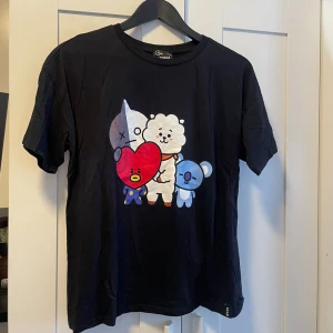 Svart BT21 t-shirt med karaktärer - Säljer en svart t-shirt från BT21 med färgglada karaktärer både fram och bak. T-shirten är i storlek L och har korta ärmar. Perfekt för fans av BT21 och deras söta figurer! 🖤 Oversize 